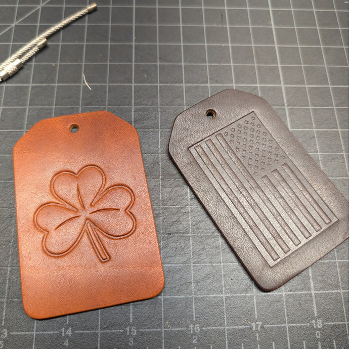 Two leather tags, one with a clover design and the other with an American flag pattern, on a cutting mat.